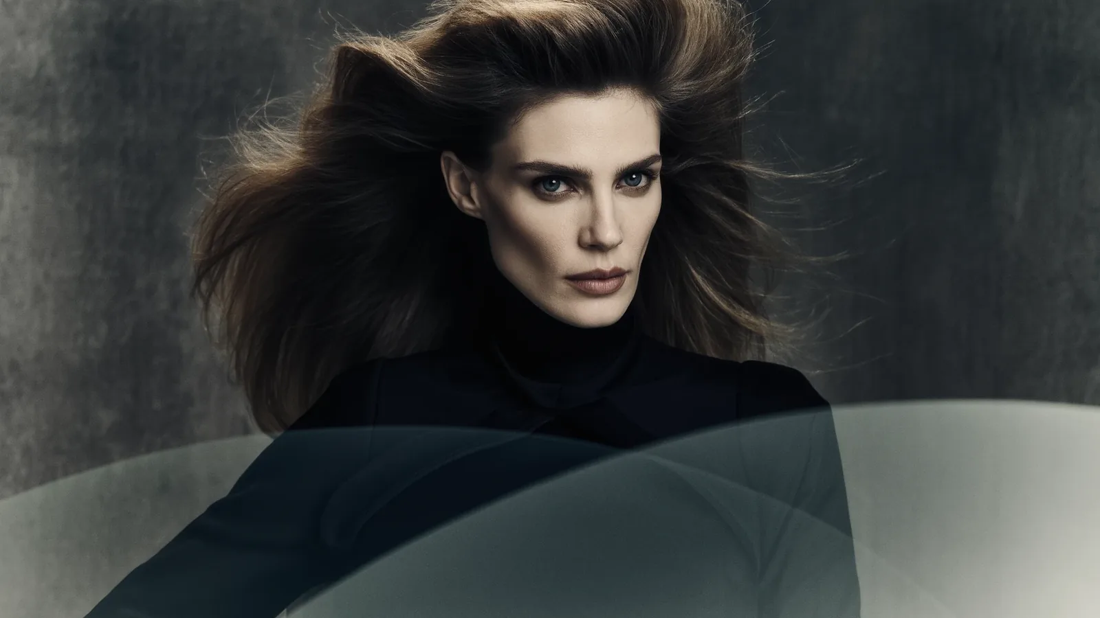 Retrato de una mujer con melena castaña al viento y jersey negro de cuello alto sobre fondo gris