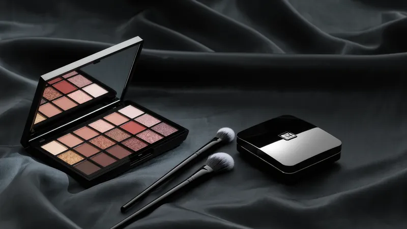 Paleta de sombras abierta con espejo, dos pinceles y una polvera negra sobre tela oscura